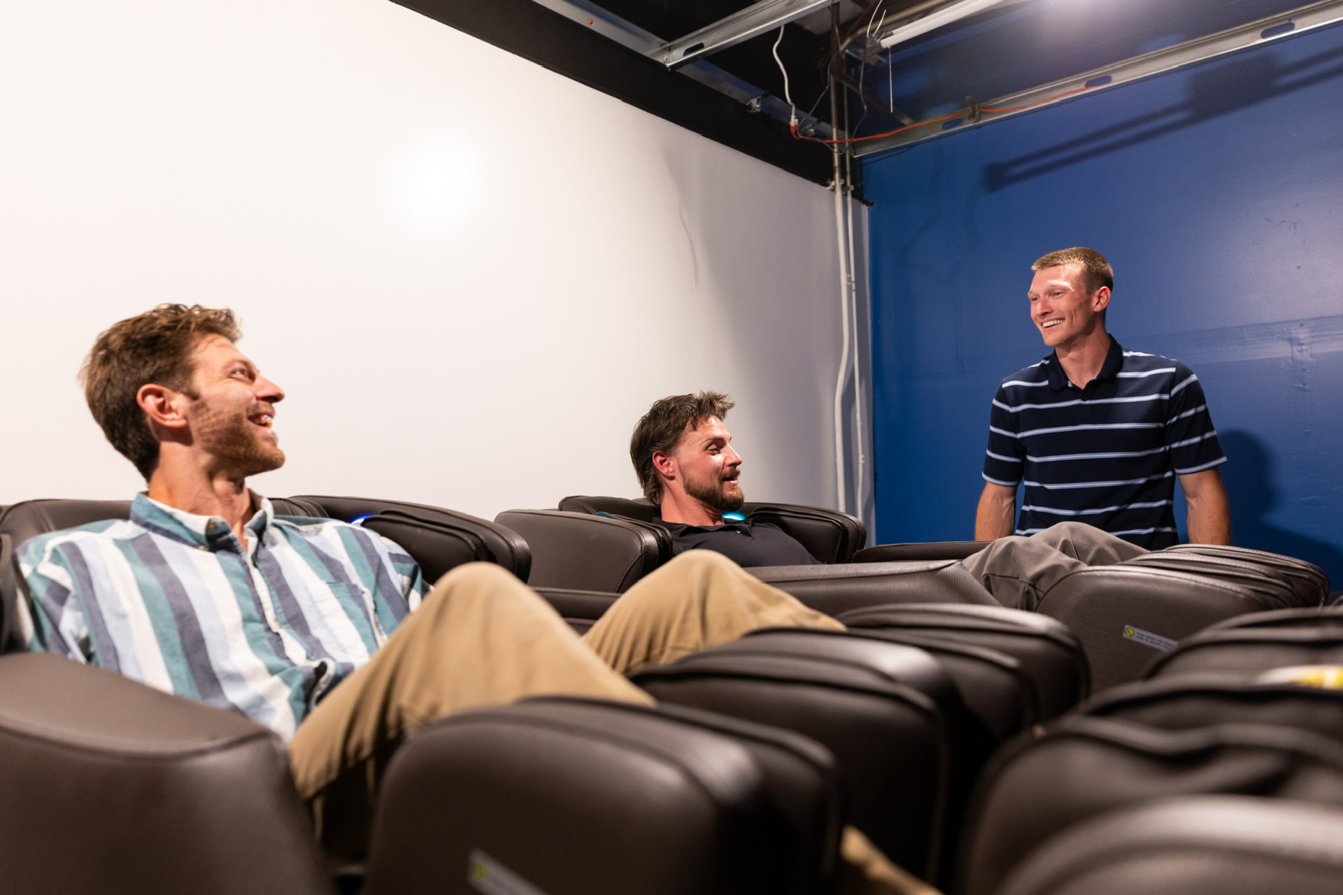 Two coworkers laughing in massage chairs while a colleague chats with them in an office break room